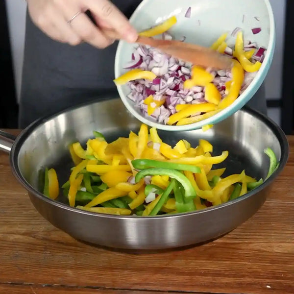 Model is adding chopped bell pepper to the sliced onion in a pan.