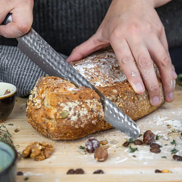 Two hands are holding the KOTAI's Serrated Knife to cut a bread displayed on the KOTAI's bamboo cutting board. Some ingredients such as flour, butter, nuts, etc. are also displayed around the bread