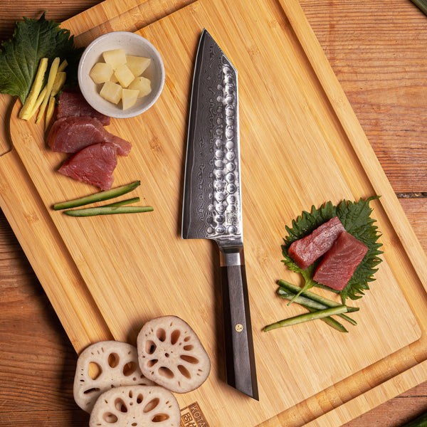 KOTAI's Santoku BUNKA Collection in Damascus steel, displayed on the KOTAI's bamboo cutting board with some ingredients displayed on it; herbs, fish, etc.