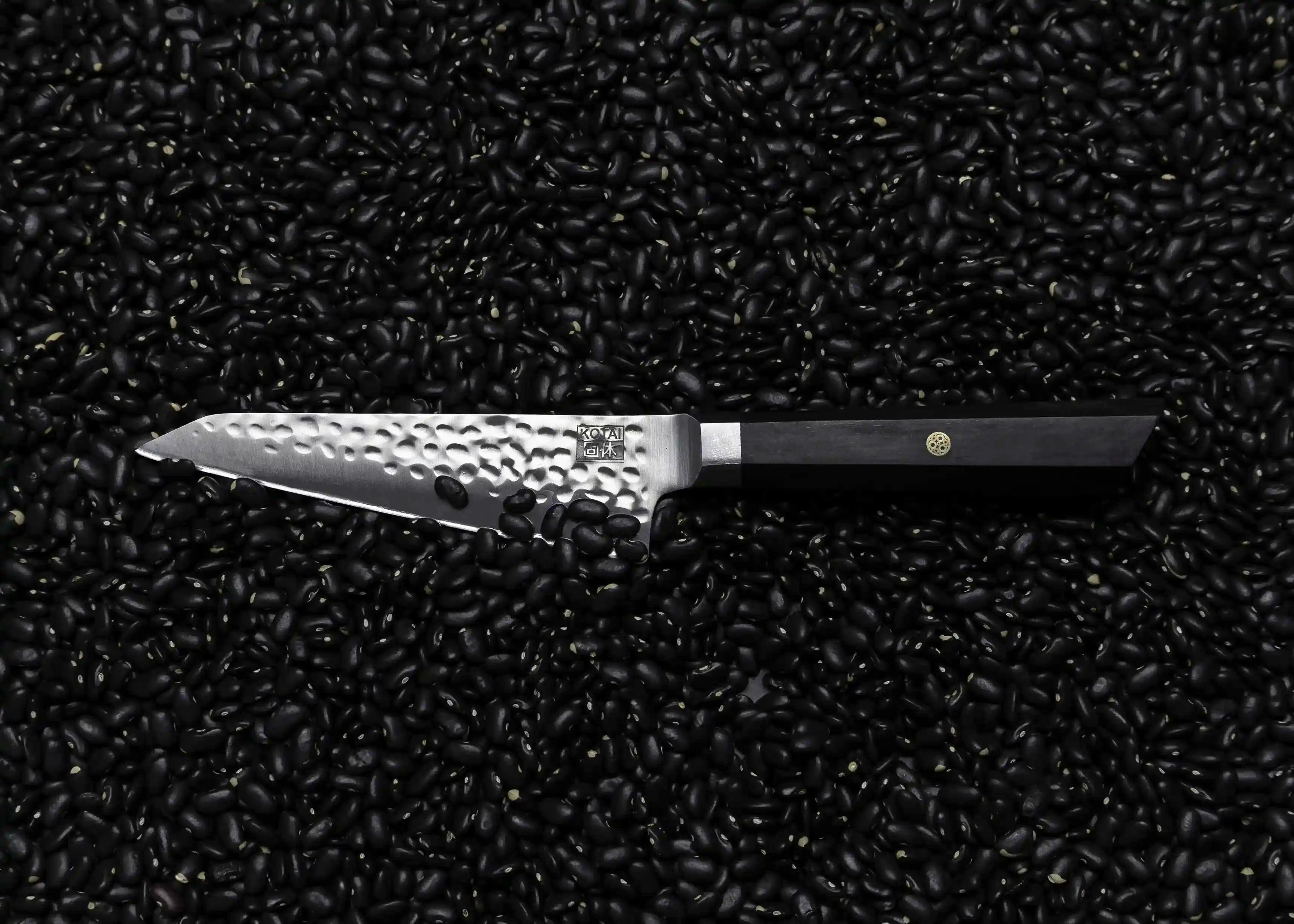 The KOTAI's Petty knife from the BUNKA Collection, horizontally displayed on a black beans background.