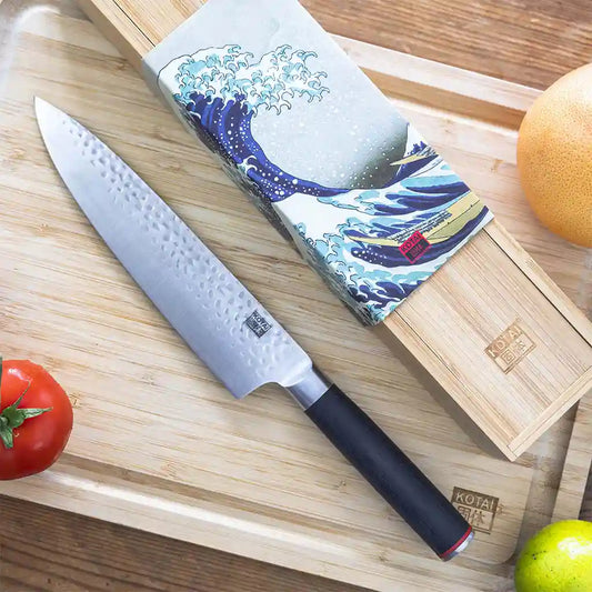 The KOTAI's Gyuto on the KOTAI's bamboo cutting board, next to its box in bamboo too with its sleeve representing the Great Wave off Kanagawa. Vegetables can be seen at the extremities of the picture.