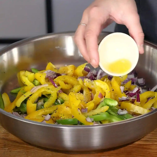 Model is adding olive oil to the mixture of chopped bell pepper and sliced onions. 