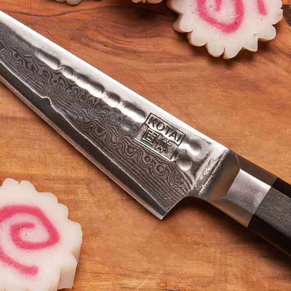 zoom on the kotai paring knife from the bunka collection to appreciate the wavy design created by the damascus steel, displayed on a wood cutting board next to narutomaki (fish surimi white and pink)