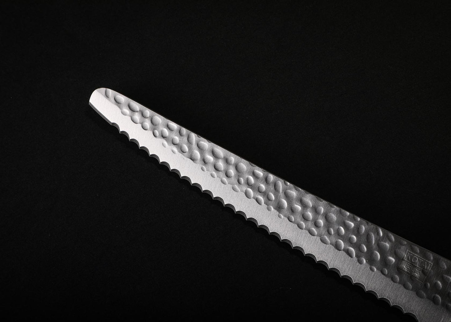 KOTAI serrated bread knife 200 mm hammered high-carbon steel blade tip on black background.